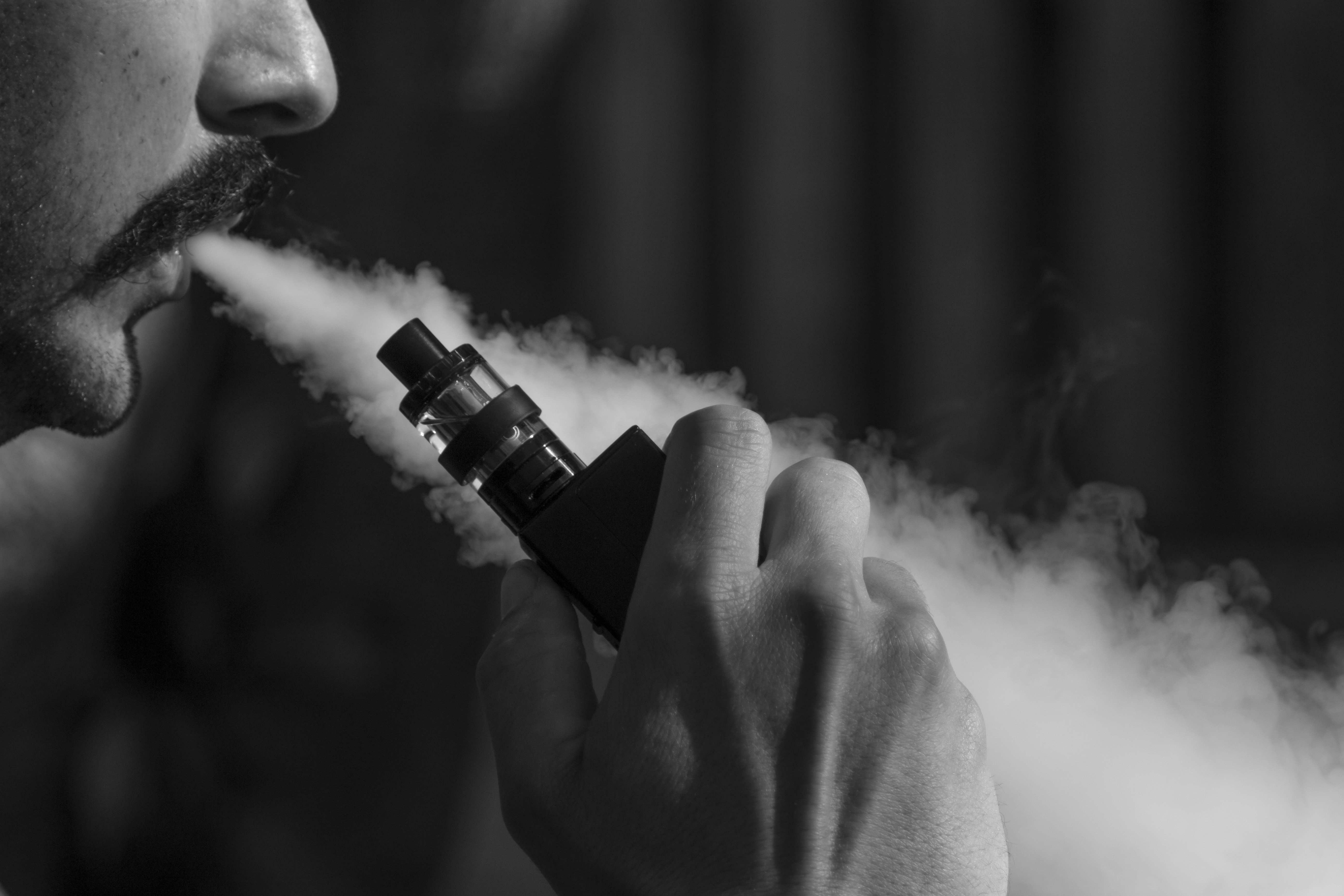 closeup profile of person with mustache exhaling while holding a vape